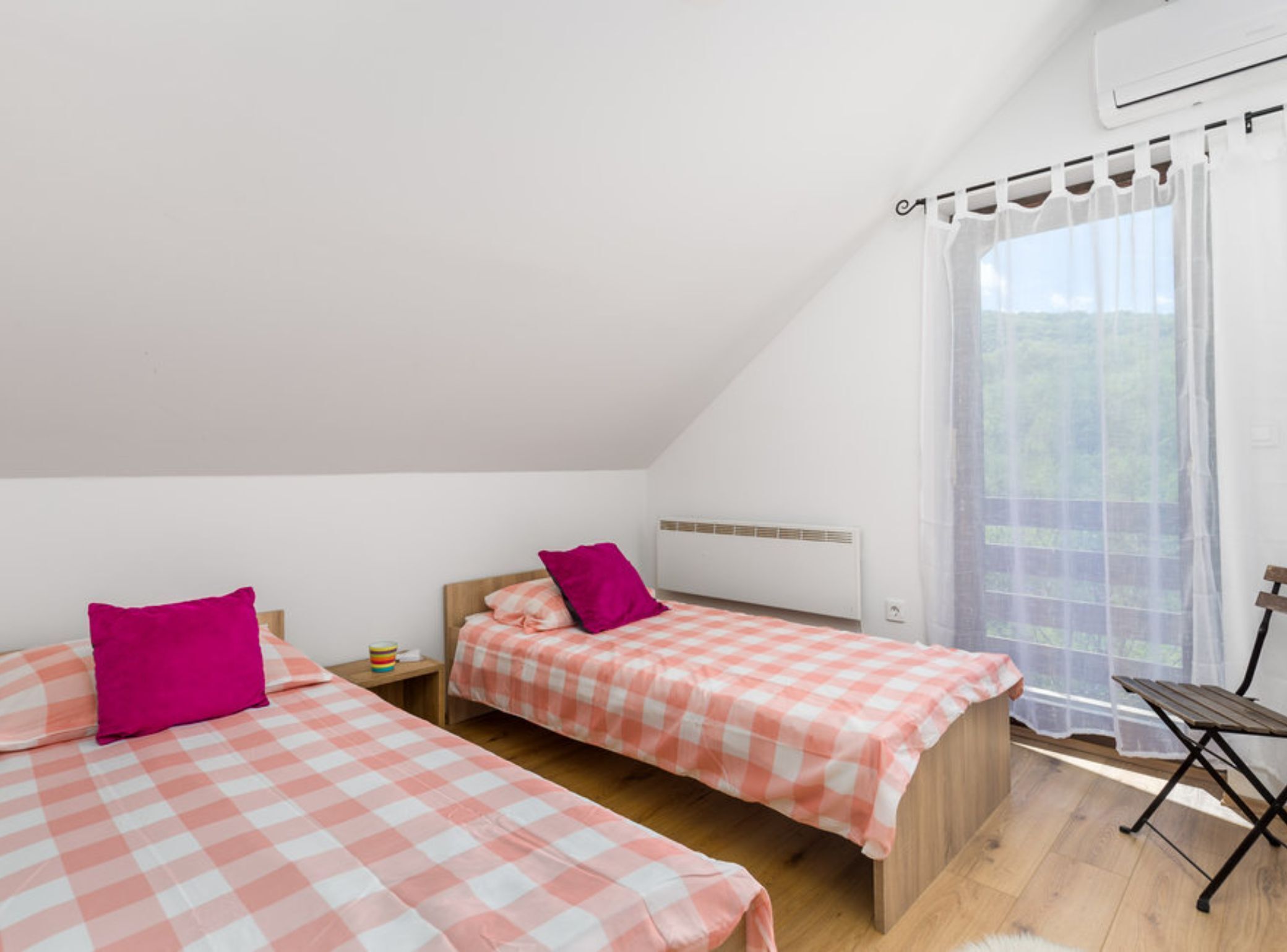 Une chambre de dortoir avec deux lits, chacun recouvert d’une couverture à carreaux rose et blanche, et orné d’un seul oreiller rose. On y trouve une petite table en bois avec une tasse colorée, un radiateur blanc et une fenêtre avec des rideaux légers.