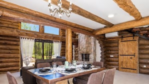 A log cabin interior with a dining table set for four, a stone fireplace, and a large window overlooking greenery. - Holiday Home Elizabeta (Slunj)