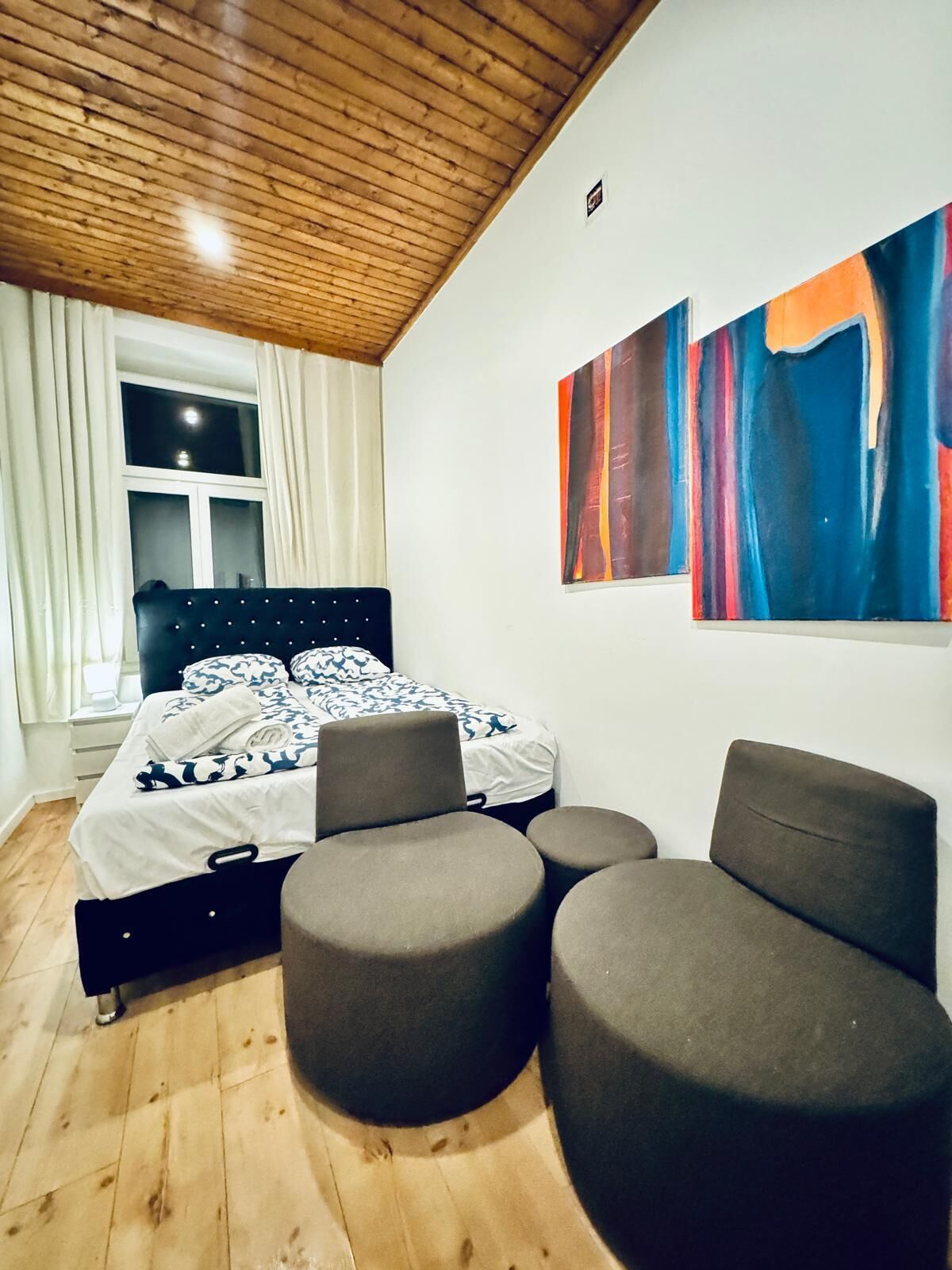 Une chambre à coucher avec un lit, une fenêtre avec des rideaux, un plafond en bois et une œuvre d’art abstraite au mur.