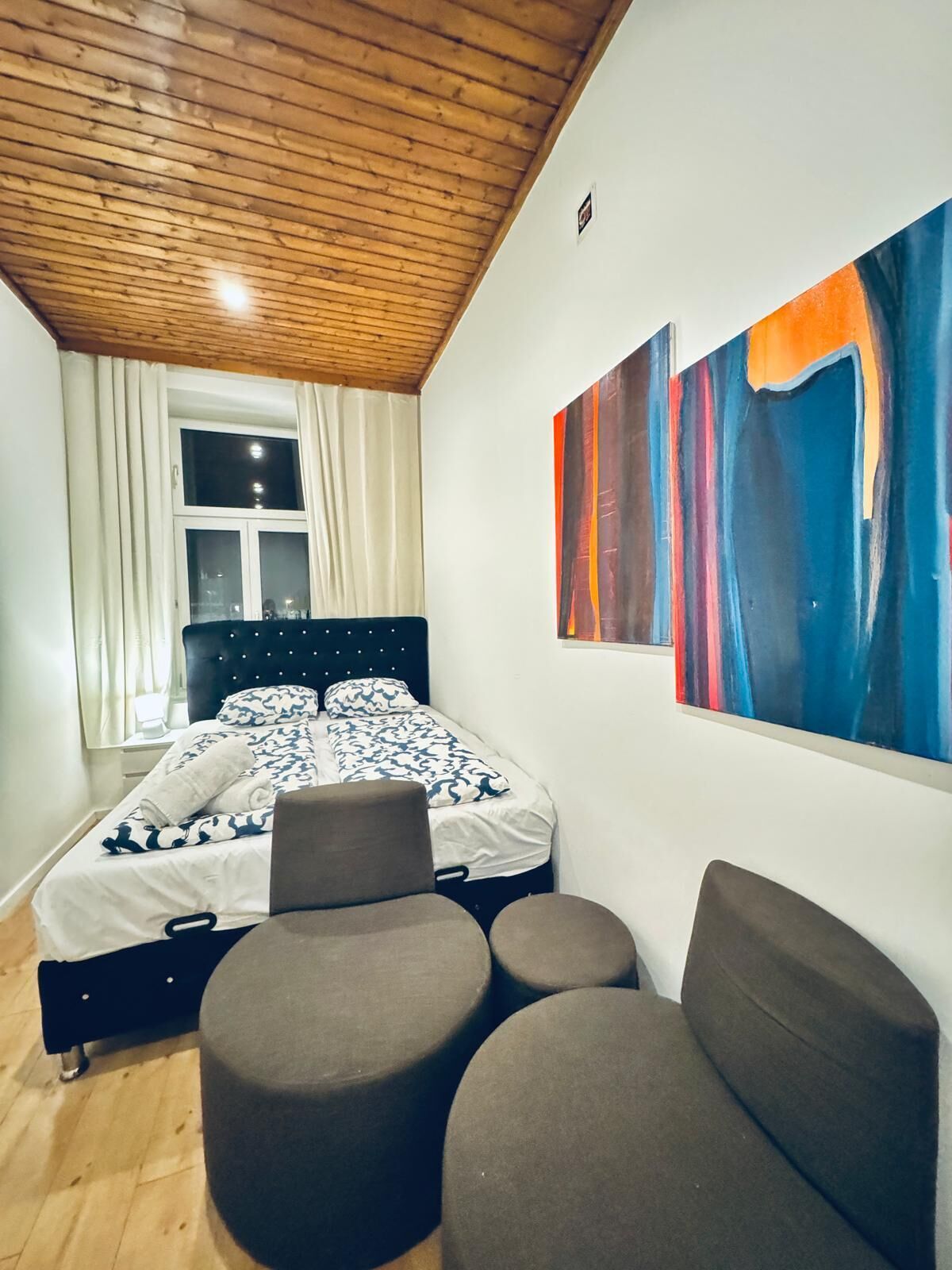 A bedroom with a bed, a large abstract painting, a window with curtains, and a wooden ceiling.