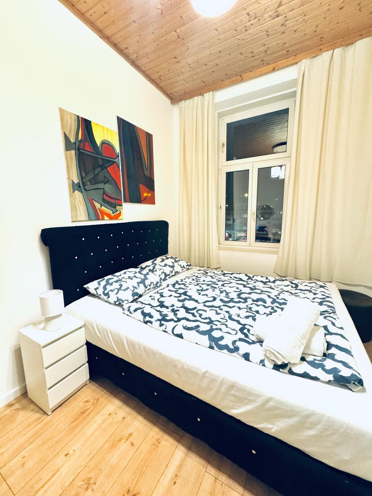 Een slaapkamer met een bed, een nachtkastje, een raam met gordijnen en twee abstracte schilderijen aan de muur.