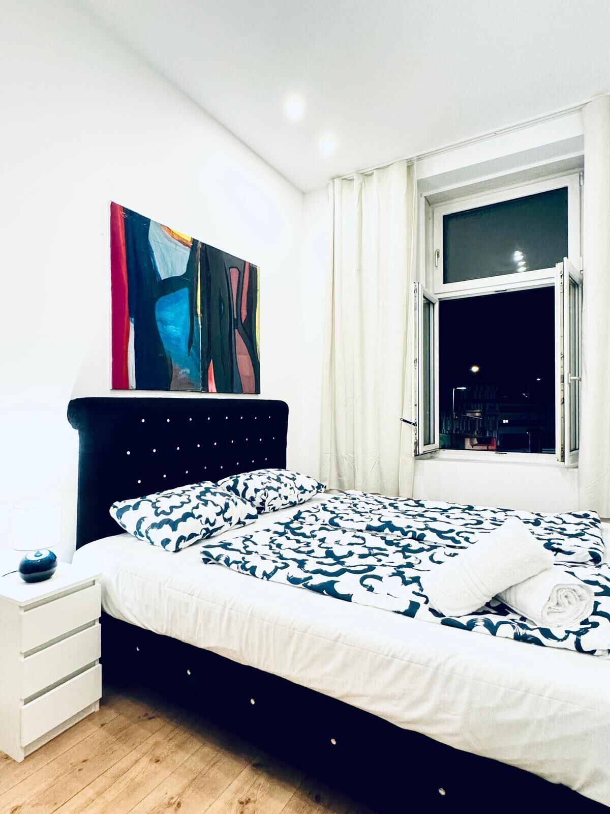 A modern bedroom with a large bed, a nightstand, a window with curtains, and a city view at night.