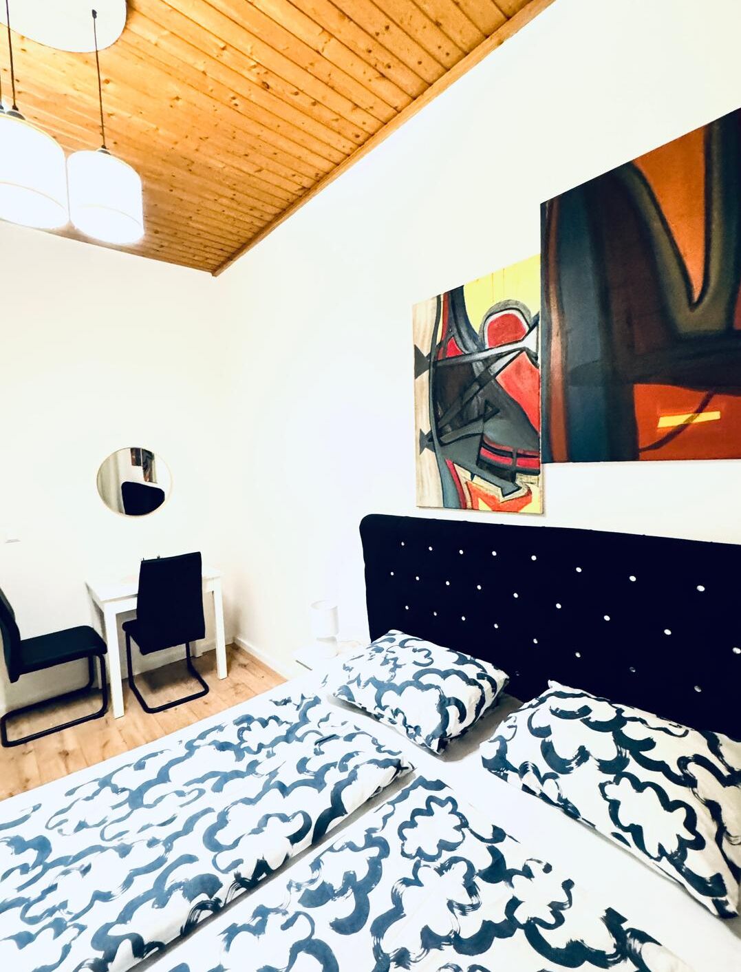 Ein Schlafzimmer mit Holzbalkendecke, einem Bett mit gemusterten Kissen, einem Schreibtisch mit Stuhl und abstrakter Kunst an der Wand.