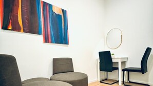 A modern room with a wooden ceiling, a white wall, and a wooden floor. There are two round grey ottomans, a small white table with a round mirror, and two black chairs. - 5BR Apartment Near Kaufpark Alterlaa (Vienna)