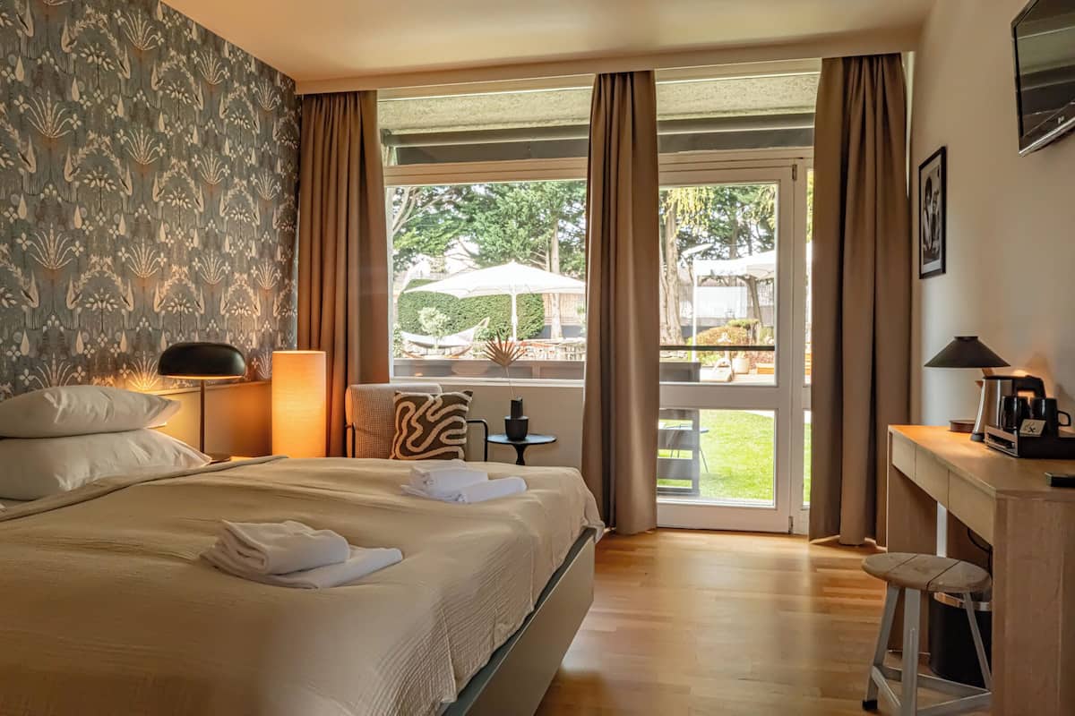 Quarto de hotel com uma cama grande, uma mesa com luminária, uma cadeira e vista para um pátio externo com mesa e guarda-sol.