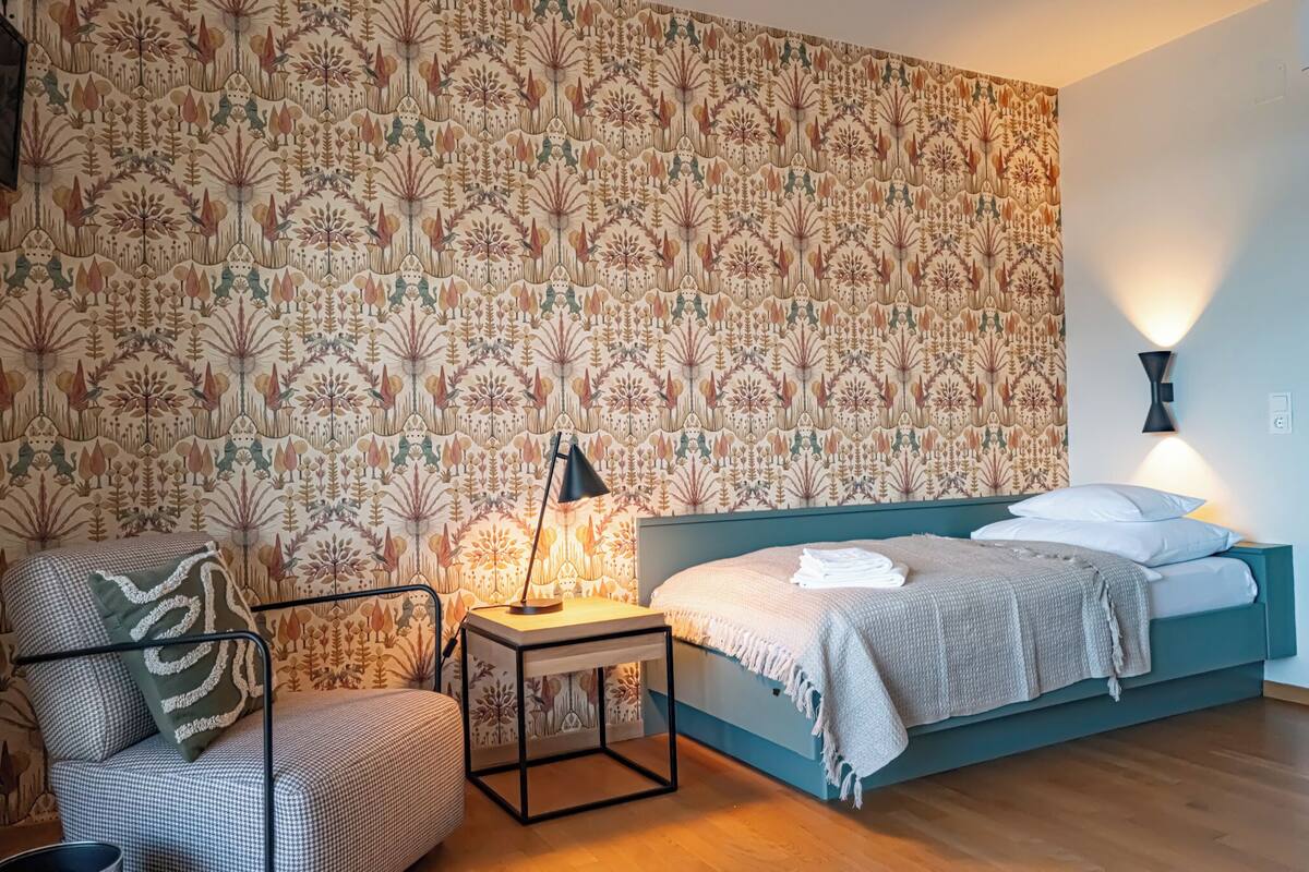 Um quarto com papel de parede estampado, uma cama com manta branca, uma cadeira com almofada estampada e uma mesa de cabeceira com abajur.