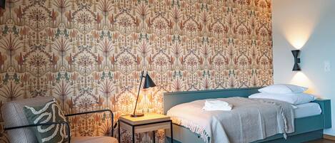 Une chambre avec un papier peint à motifs, un lit recouvert d’une couverture blanche, une chaise agrémentée d’un coussin à motifs et une table de chevet avec une lampe.