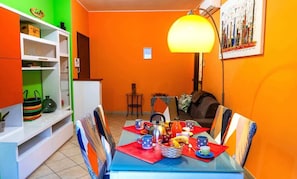 A dining area with a table set for two, chairs, a lamp, and a shelf with kitchen items.