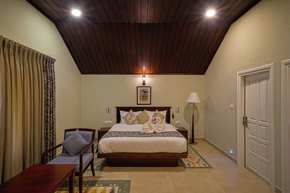A bedroom with a wooden ceiling, a bed with decorative pillows, a bedside table, a chair, a lamp, and a door.