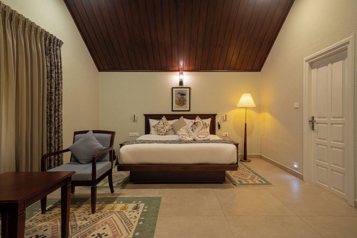 A bedroom with a wooden ceiling, a bed with pillows, a chair, a lamp, and a door.