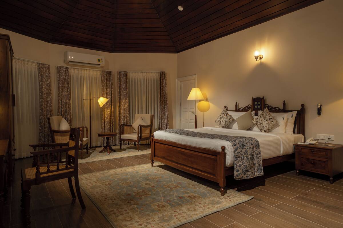A bedroom with a wooden bed, two bedside lamps, a nightstand, a chair, a desk, and an air conditioning unit.