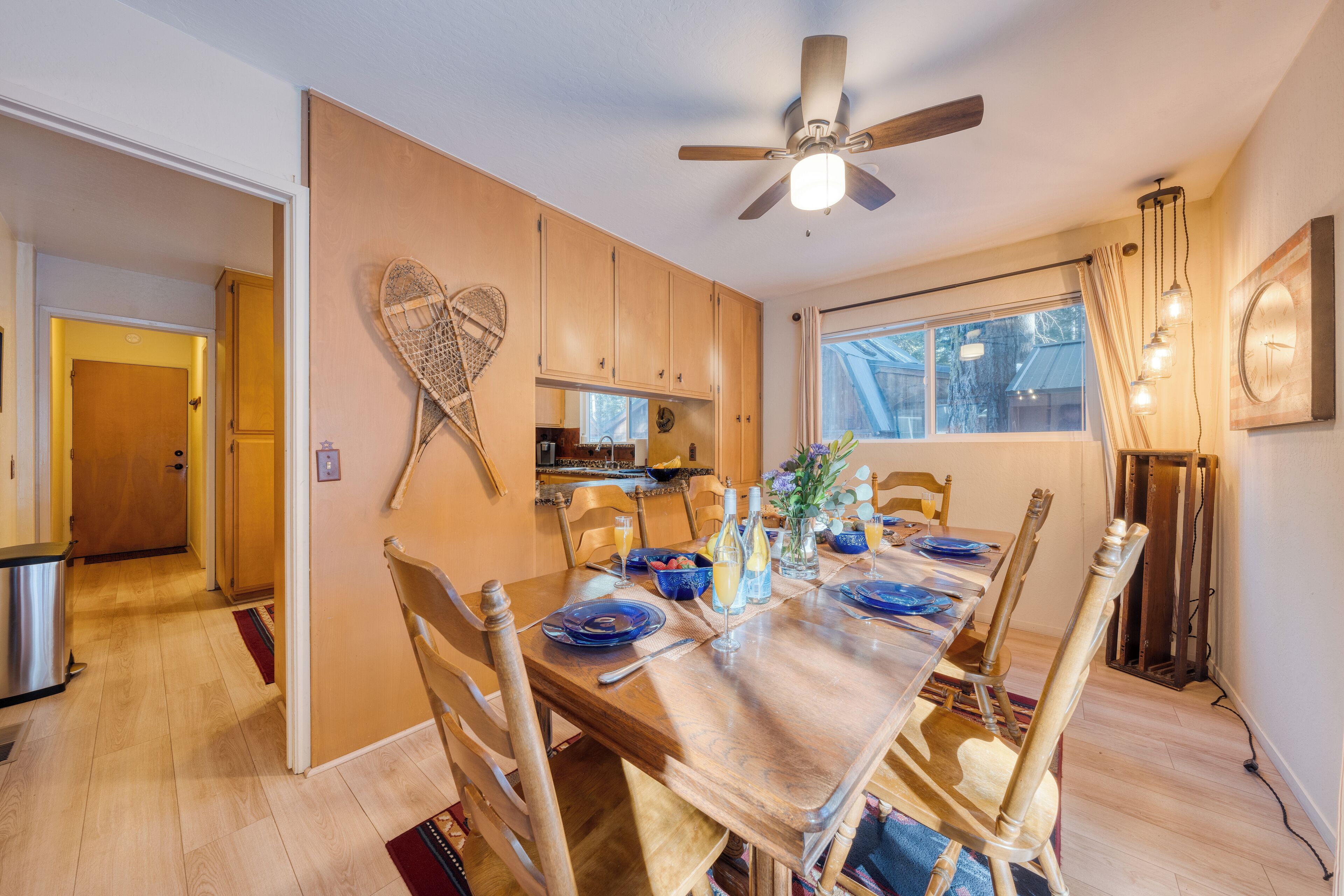A dining area with a wooden table set for six, surrounded by wooden chairs. A ceiling fan is above the table, and there is a window with curtains to the right.