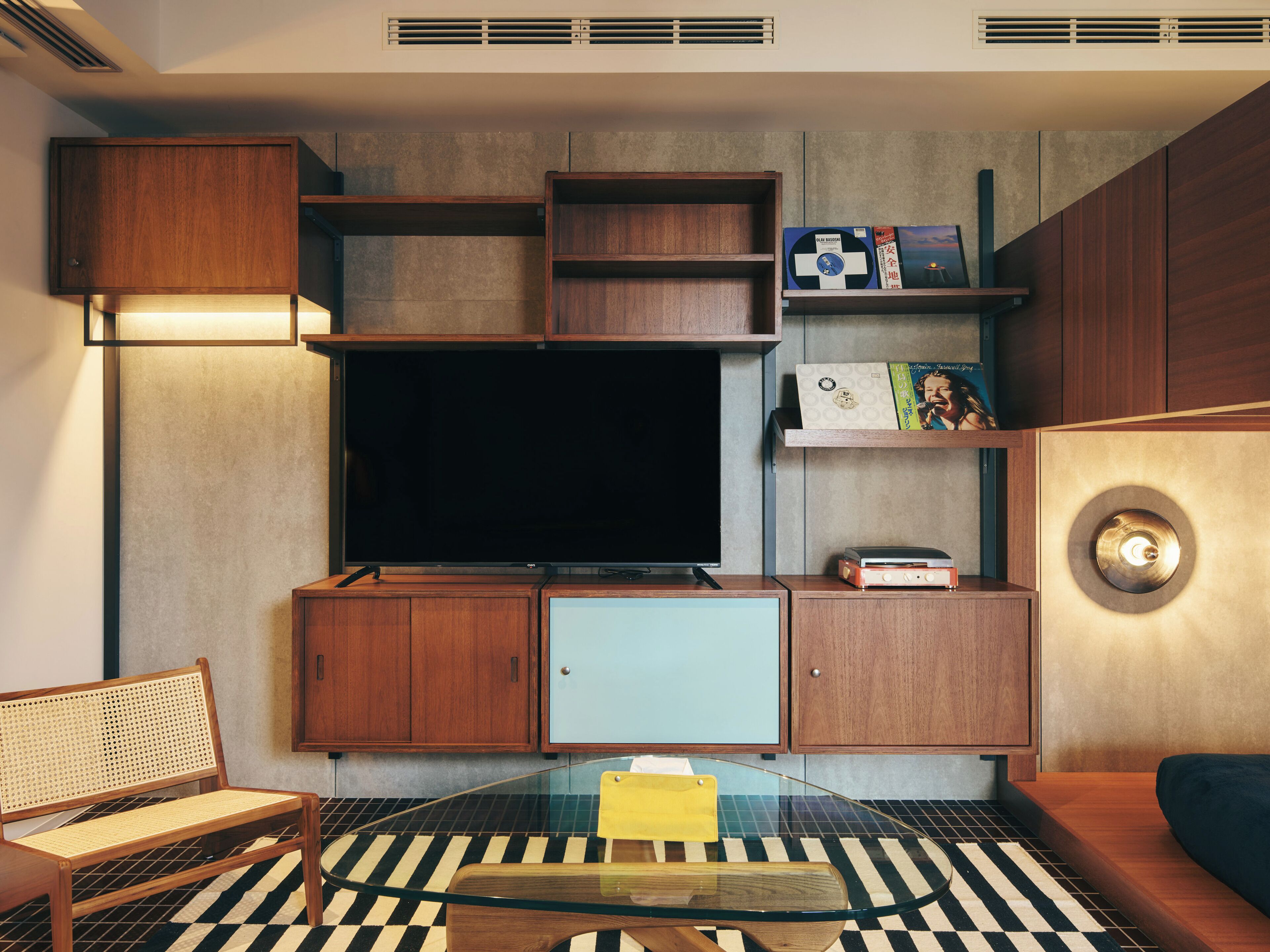 A modern living room with a flat-screen TV, wooden cabinets, a glass coffee table, and a checkered floor.