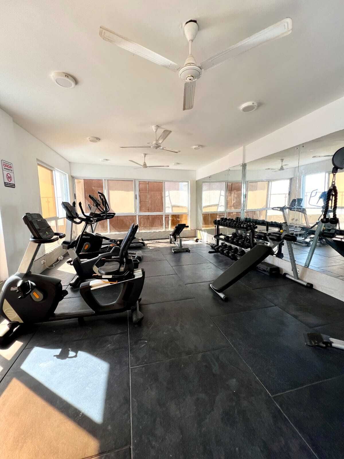 Une salle de sport équipée de nombreux appareils d’exercice, notamment des tapis de course et des bancs de musculation, ainsi que de ventilateurs au plafond.