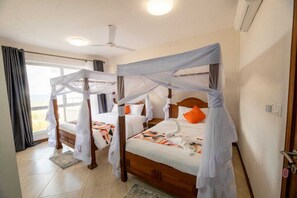 A room with two wooden beds, each with a canopy and white bedding. There is a ceiling fan, a window with curtains, and a door.