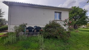 A single-story building with a white exterior, a small window, and a door. There is a metal gate in front, and the surrounding area is lush with greenery and flowering plants. - Quiet, 70s-style house with large garden in the heart of the Lot et Garonne region (Saint-Étienne-de-Fougères)