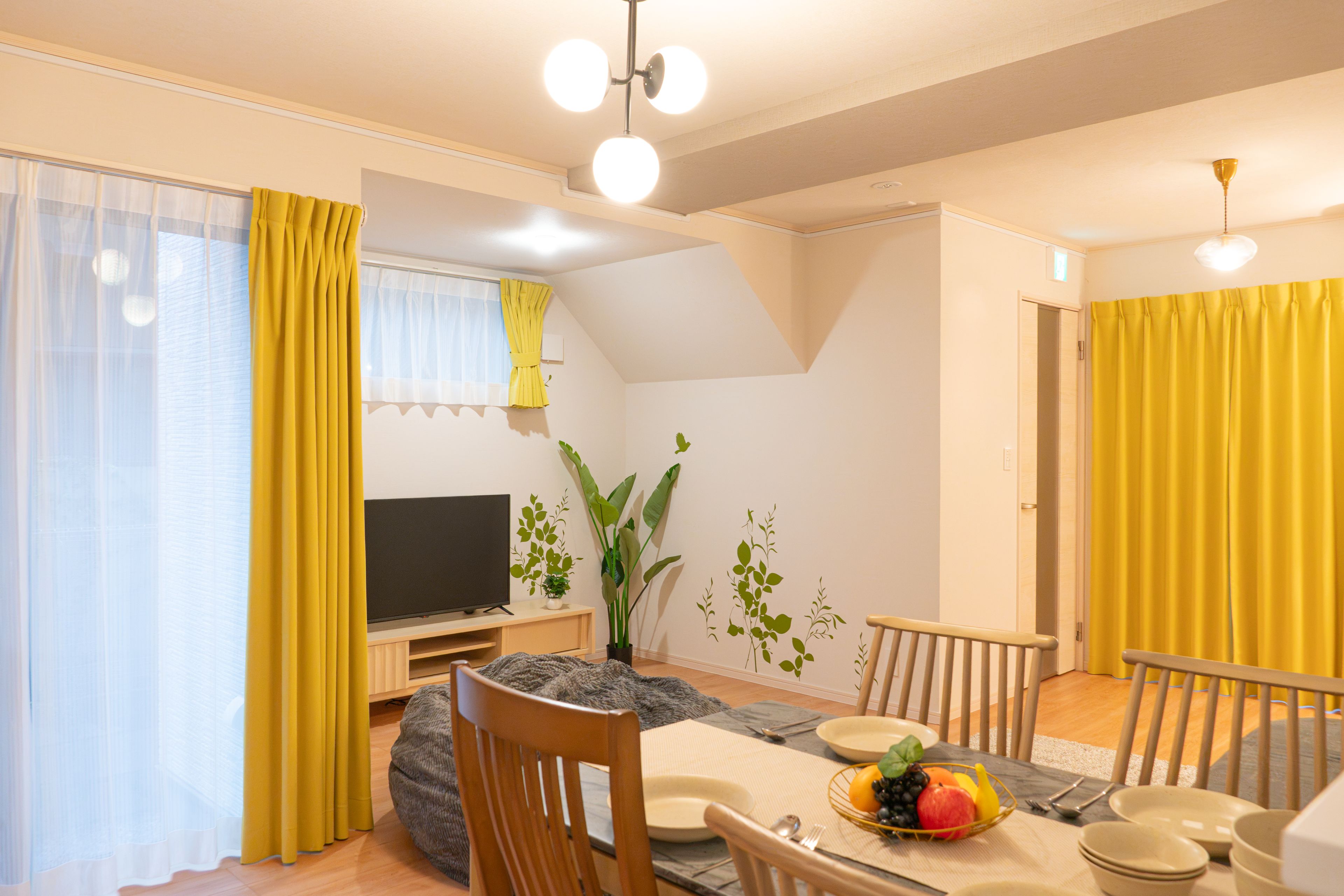 A dining area with a table set for four, a television on a stand, and a plant on the wall.