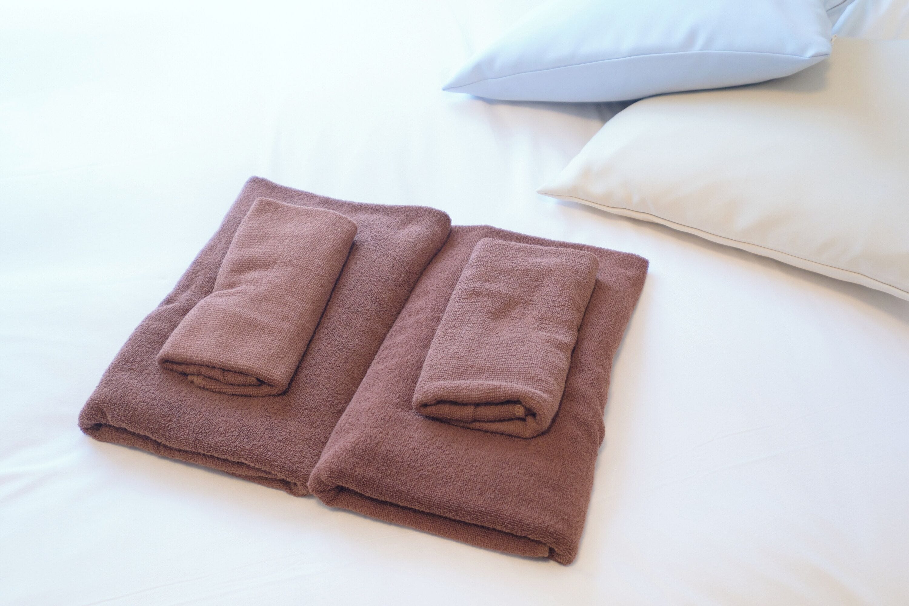 Two neatly folded brown towels on a bed with white bedding and a light-colored pillow.