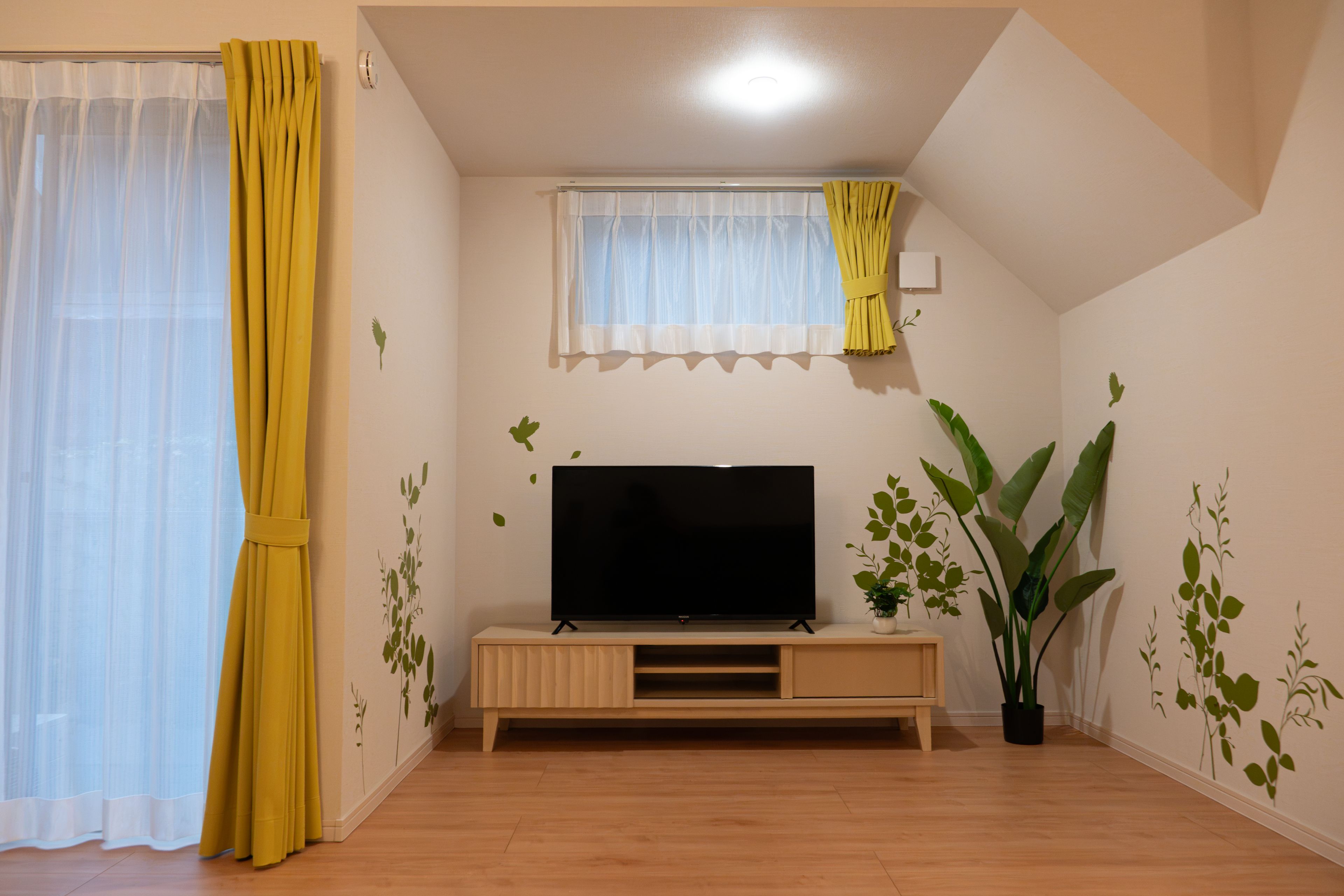 Un salon avec un téléviseur à écran plat posé sur un meuble en bois, une fenêtre ornée de rideaux légers et une plante en pot.