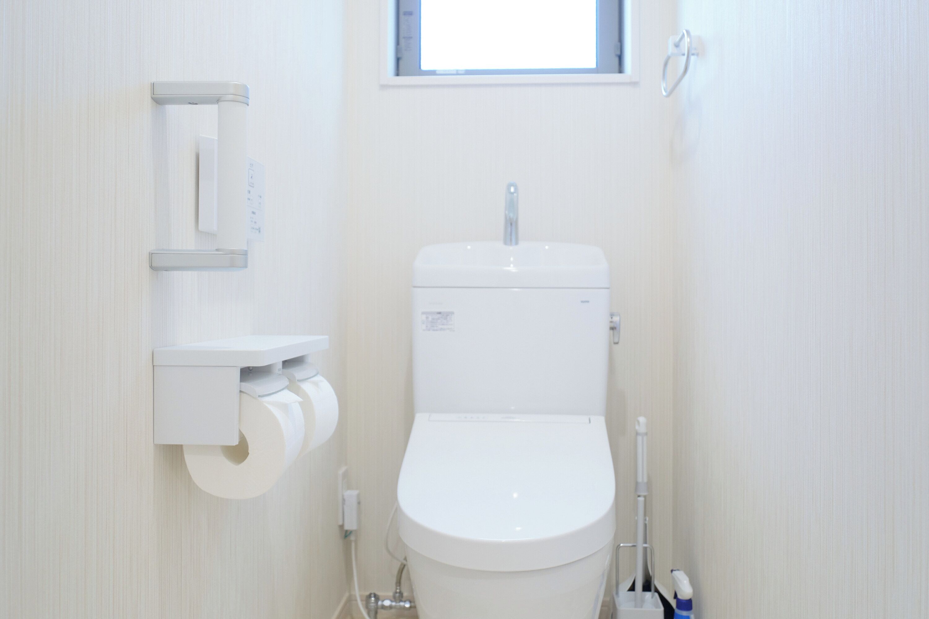 Ein kleines, weißes Badezimmer mit Toilette, einem an der Wand befestigten Toilettenpapierhalter und einem Fenster über der Toilette.