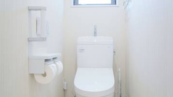 Ein kleines, weißes Badezimmer mit Toilette, einem an der Wand befestigten Toilettenpapierhalter und einem Fenster über der Toilette.