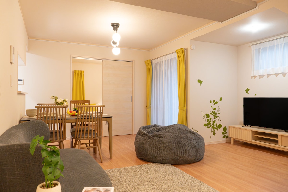 Una sala de estar con sofá, sillón de peluche, mesa de comedor con sillas, televisor y planta.