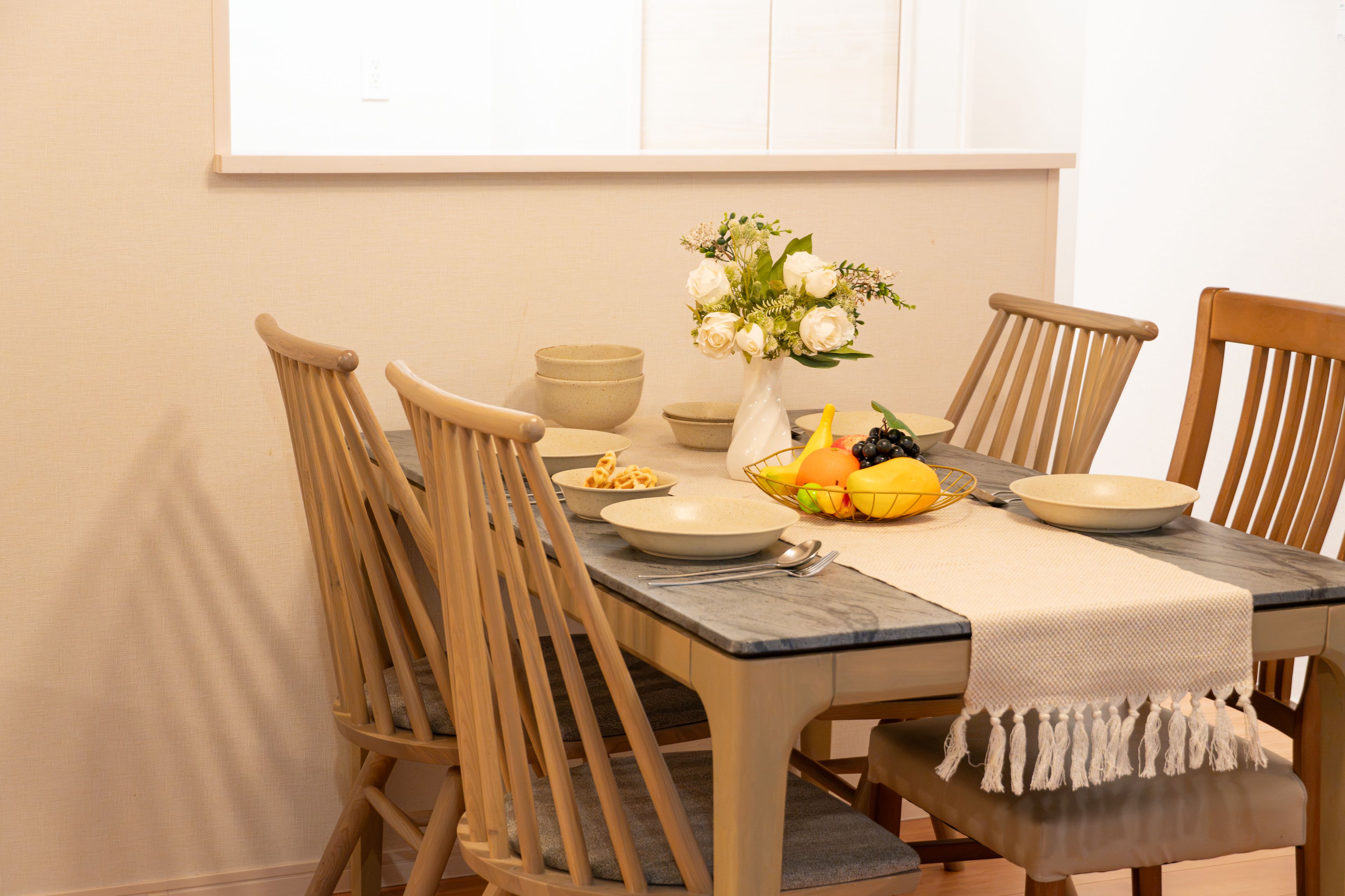 A dining table with four wooden chairs, a vase with flowers, and a fruit basket.