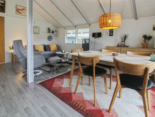 A modern living room with a dining area, featuring a large dining table, wooden chairs, a grey sofa, and a red patterned rug.