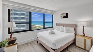 A hotel room with a large window offering a view of the beach and ocean, a neatly made bed with white linens, a bedside table with a lamp, and a small bedside table with a lamp. - Classic Resort Amenities Condo Steps from the Beach with Gym & Pool 1208 (Miami Beach)
