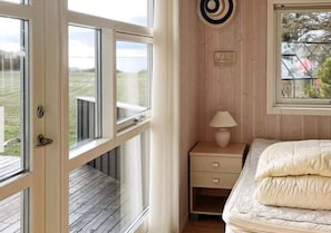 Une chambre avec un plancher en bois, un lit avec une literie blanche, une table de chevet avec une lampe, une fenĂȘtre donnant sur lâextĂ©rieur et une porte menant Ă un balcon.