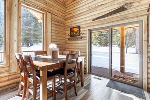 Interior de una cabaña de madera con una mesa de comedor, sillas, chimenea y un ventanal grande.