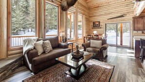 A log cabin interior with a large window, a mounted deer head, a sofa, a chair, and a dining area. - Wrangler’s Retreat | 7 Mi to YNP | Private Pond (West Yellowstone)