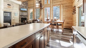 A log cabin interior with a stone fireplace, wooden dining table, and a large window with a deer head decoration. - Wrangler’s Retreat | 7 Mi to YNP | Private Pond (West Yellowstone)