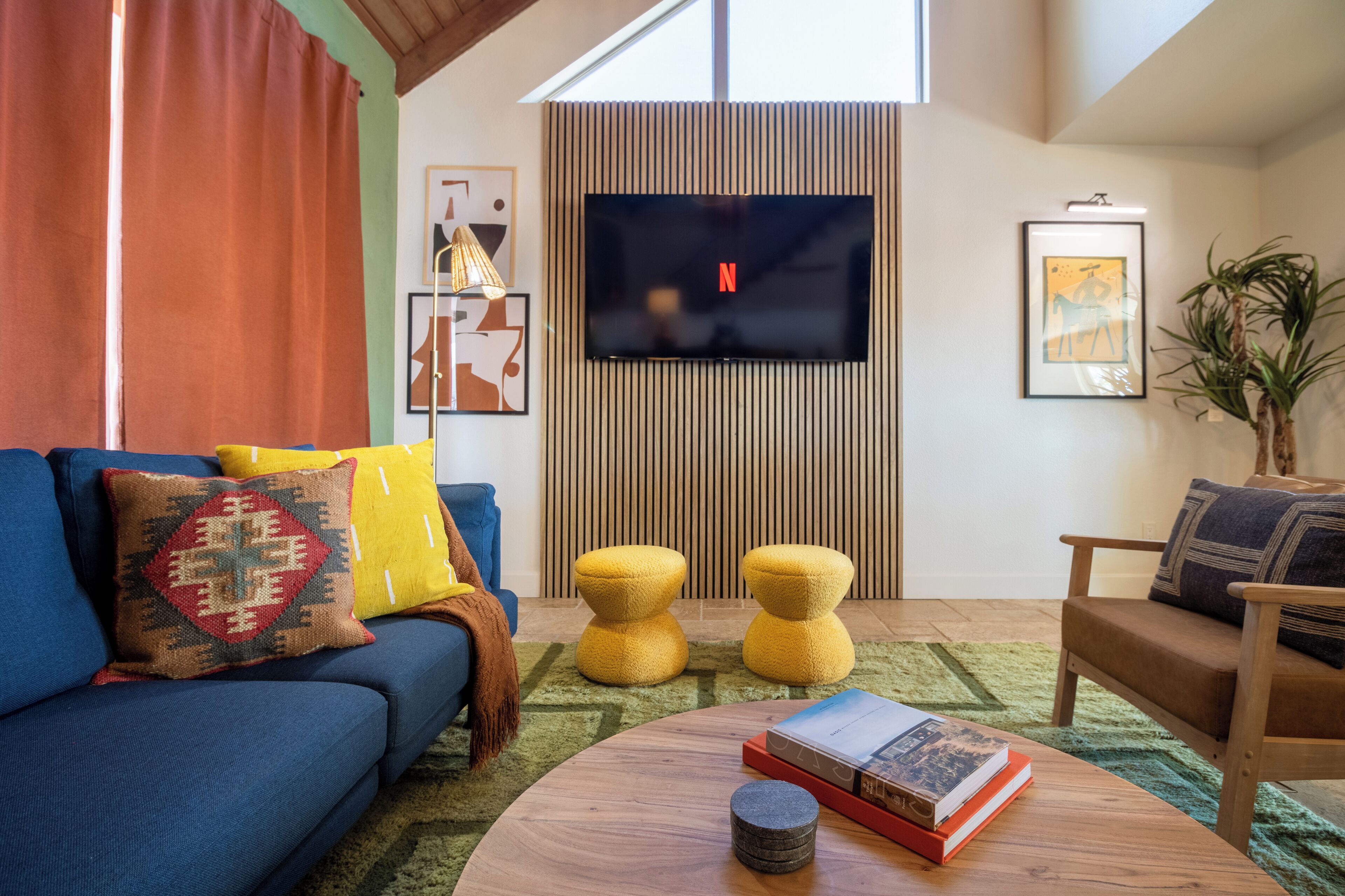 A modern living room with a blue sofa, yellow ottomans, a round coffee table with books, and a flat-screen TV mounted on a wooden panel wall.