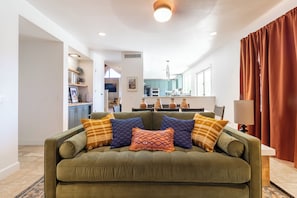 A living room with a green sofa, colorful cushions, a kitchen in the background, and a window with curtains.