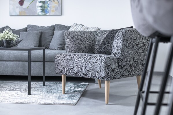Uma sala de estar moderna com um sofá cinzento, uma poltrona estampada, uma mesa de centro preta e um vaso decorativo com flores.