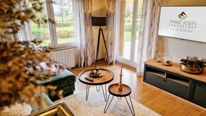Een kamer met uitzicht op een tuin, een staande lamp, een klein rond tafeltje met een afstandsbediening, een bank en een kast met decoratieve objecten.