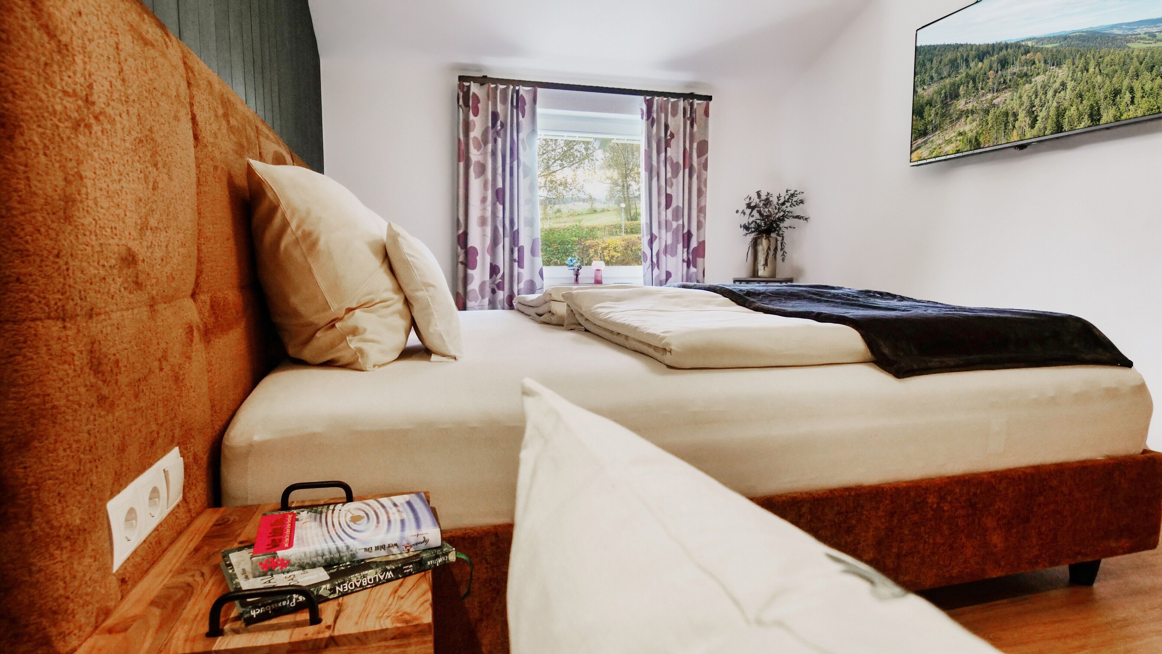 Een slaapkamer met een bed, een raam met gordijnen, uitzicht op het groen en een klein tafeltje met boeken.