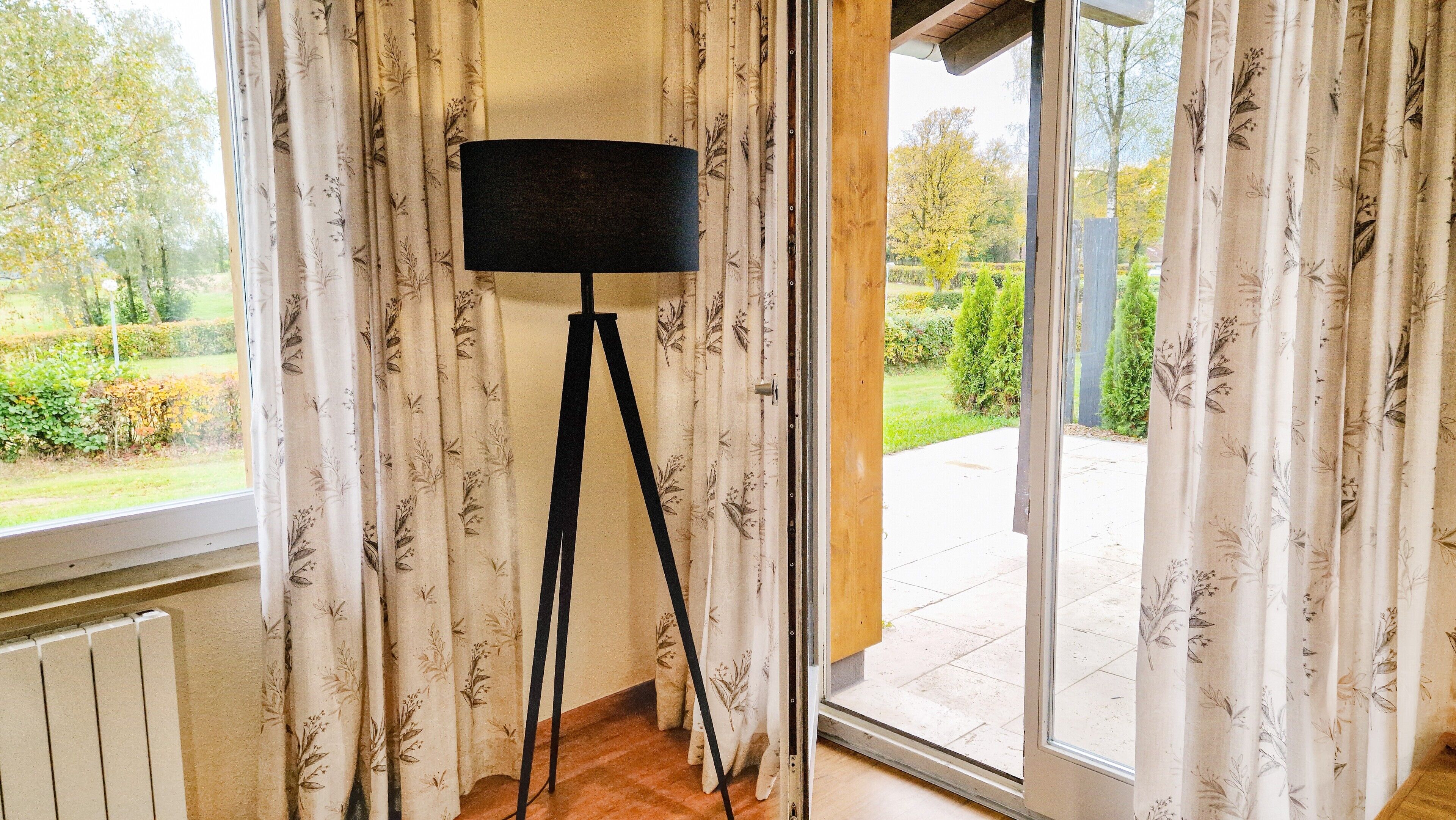 Una habitación con una lámpara de pie, una puerta corrediza de vidrio que da a un jardín y cortinas estampadas.