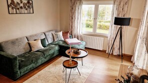 En stue med en grøn sofa, et lille rundt bord, en gulvlampe og et vindue med gardiner.