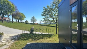 A modern building with a wooden deck, a stone sculpture, and a view of a green landscape with trees and a distant mountain. - Tiny Home 'Franzl' with Mountain View, Wi-Fi and Air Conditioning (Untergriesbach)