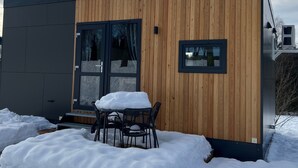 A modern wooden cabin with a glass door, a small table with chairs, and a snow-covered ground. - Tiny Home 'Franzl' with Mountain View, Wi-Fi and Air Conditioning (Untergriesbach)