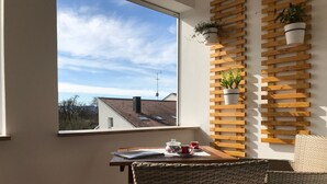 A balcony with a view of a tiled roof, a wooden wall-mounted plant shelf, and a table with a chair. - Apartment 'Maisonette Am See' with Lake View, Wi-Fi and Air Conditioning (Nonnenhorn)