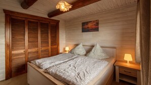 A bedroom with a wooden bed, a nightstand with a lamp, a wooden wardrobe, and a painting on the wall. - Cozy Log Cabin in the Harz with Private Terrace & Garden (Bad Sachsa)