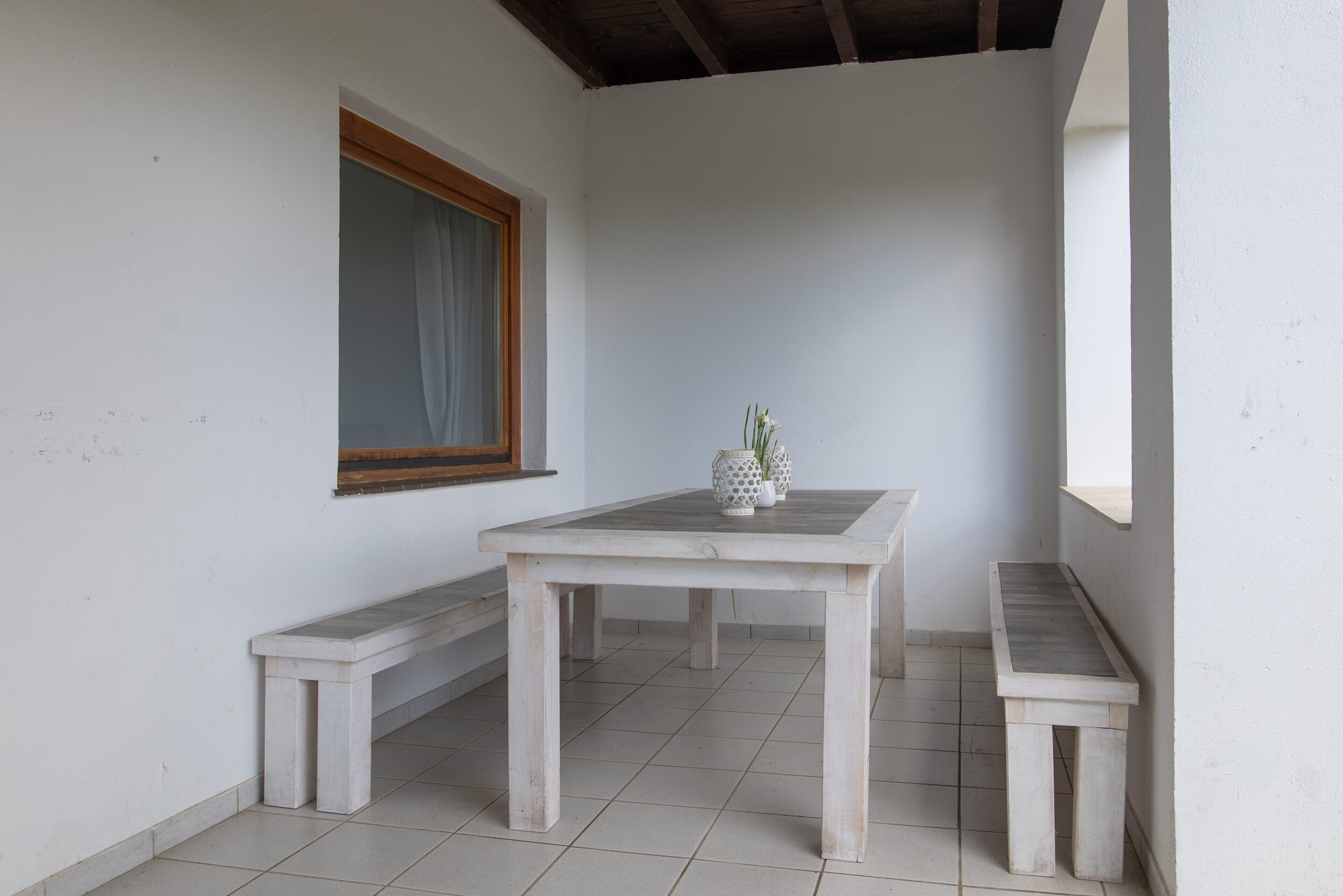 Um quarto com chão de azulejos brancos, um banco de madeira, uma mesa branca com um vaso e uma janela com moldura de madeira.