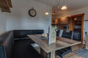 Uma área de refeições moderna com mesa de madeira, assentos em couro preto, iluminação pendente e uma cozinha visível ao fundo.