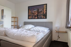 Cama bem arrumada com roupa de cama branca, cabeceira cinzenta, prateleira de madeira com abajur e pintura de paisagem urbana emoldurada na parede.