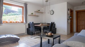 A modern living room with a sofa, a dining table with chairs, a clock on the wall, and a view of the outdoors through a window. - Cozy Log Cabin in the Harz with Private Terrace & Garden (Bad Sachsa)