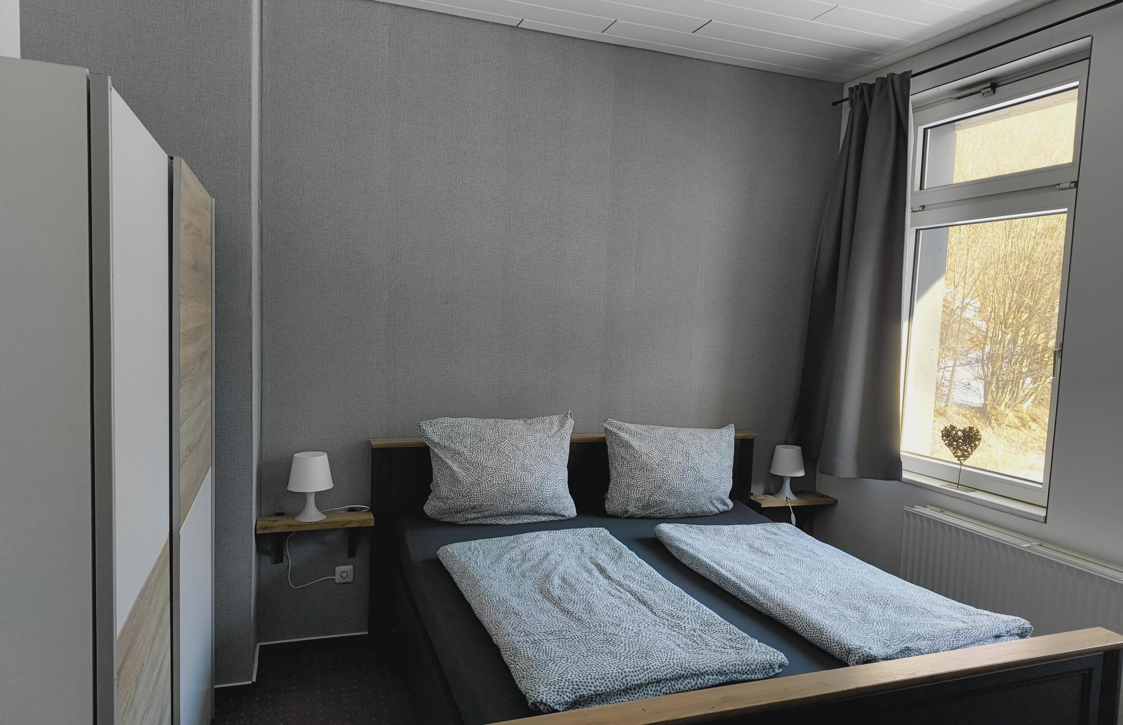 A room with a bed, two bedside tables, a window with curtains, and a radiator.