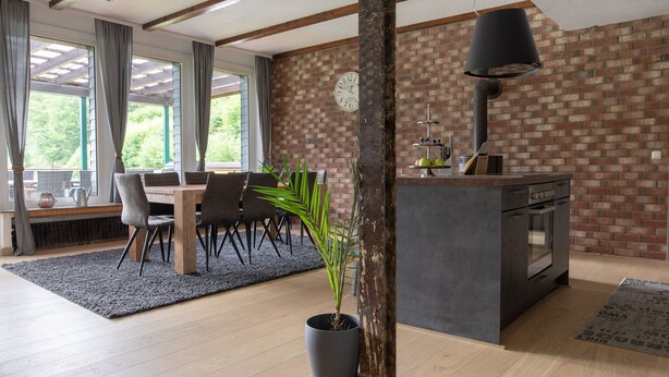 A modern kitchen with a brick wall, wooden beams, and a dining area with a table and chairs. - Holiday Home 'Beautiful View' in the Harz with Mountain View, Private Terrace and Wi-Fi (Walkenried)